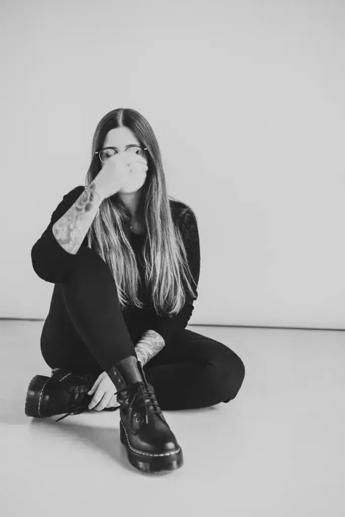 Janine mit Tattoos und Brille sitzt auf dem Boden